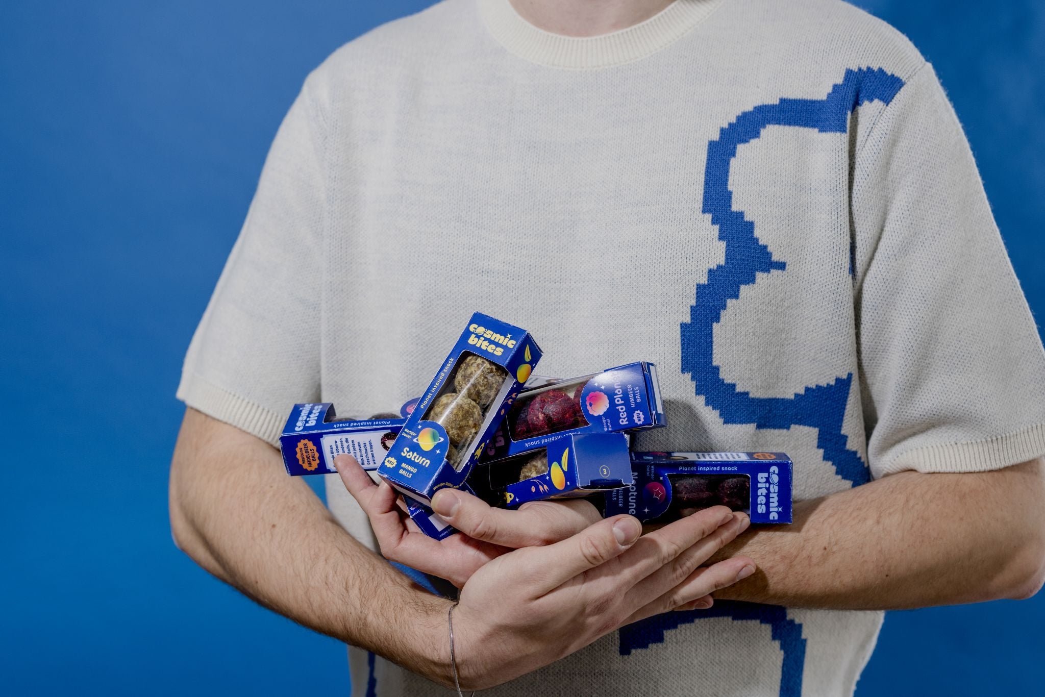Eine Handvoll galaktischer Abenteuer: Person hält verschiedene Cosmic Bites Energy Ball Packungen vor blauem Hintergrund: Himbeer-Mars, Mango-Saturn und Heidelbeer-Neptun – der ideale gesunde Snack für einen aktiven Lebensstil wird in all seinen Varianten präsentiert.