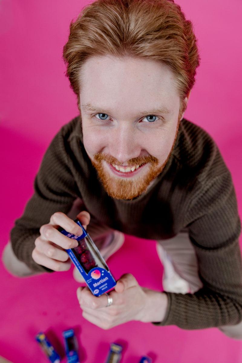 Ein junger Mann in der Hocke wird von oben fotografiert. Er schaut spielerisch hoch in die Kamera und hält mit beiden Händen eine Packung Cosmic Bites Energy Balls der Sorte Himbeere. Die strahlend roten Energy Balls, die eher wie der Planet Mars aussehen, stehen im starken Kontrast zu seinen tiefblauen Augen.