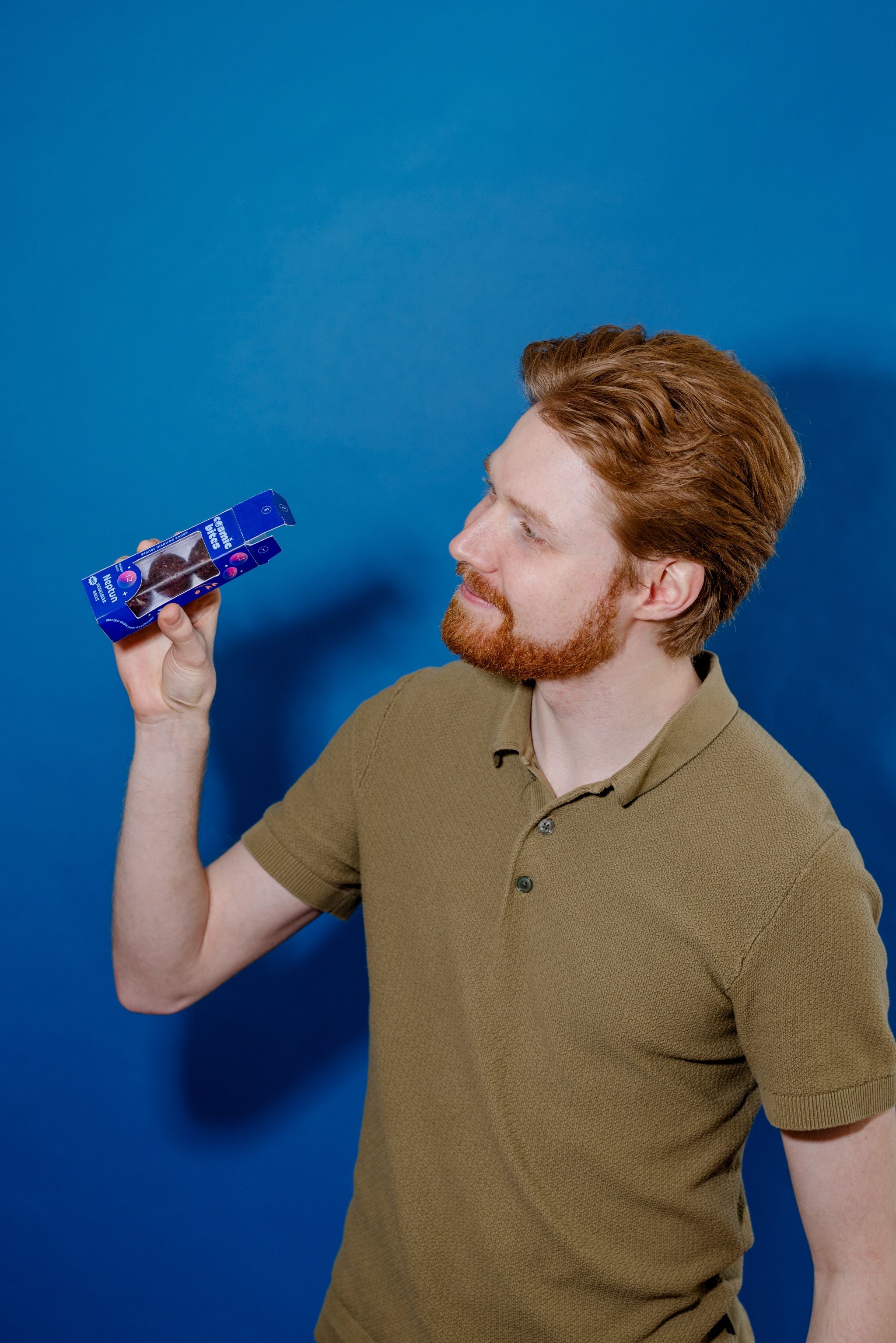 Ein junger Mann schaut spielerisch in eine geöffnete Cosmic Bites Energy Ball Packung der Sorte Heidelbeere. Jeder der tiefvioletten Bälle sieht aus wie ein kleiner Planet Neptun.