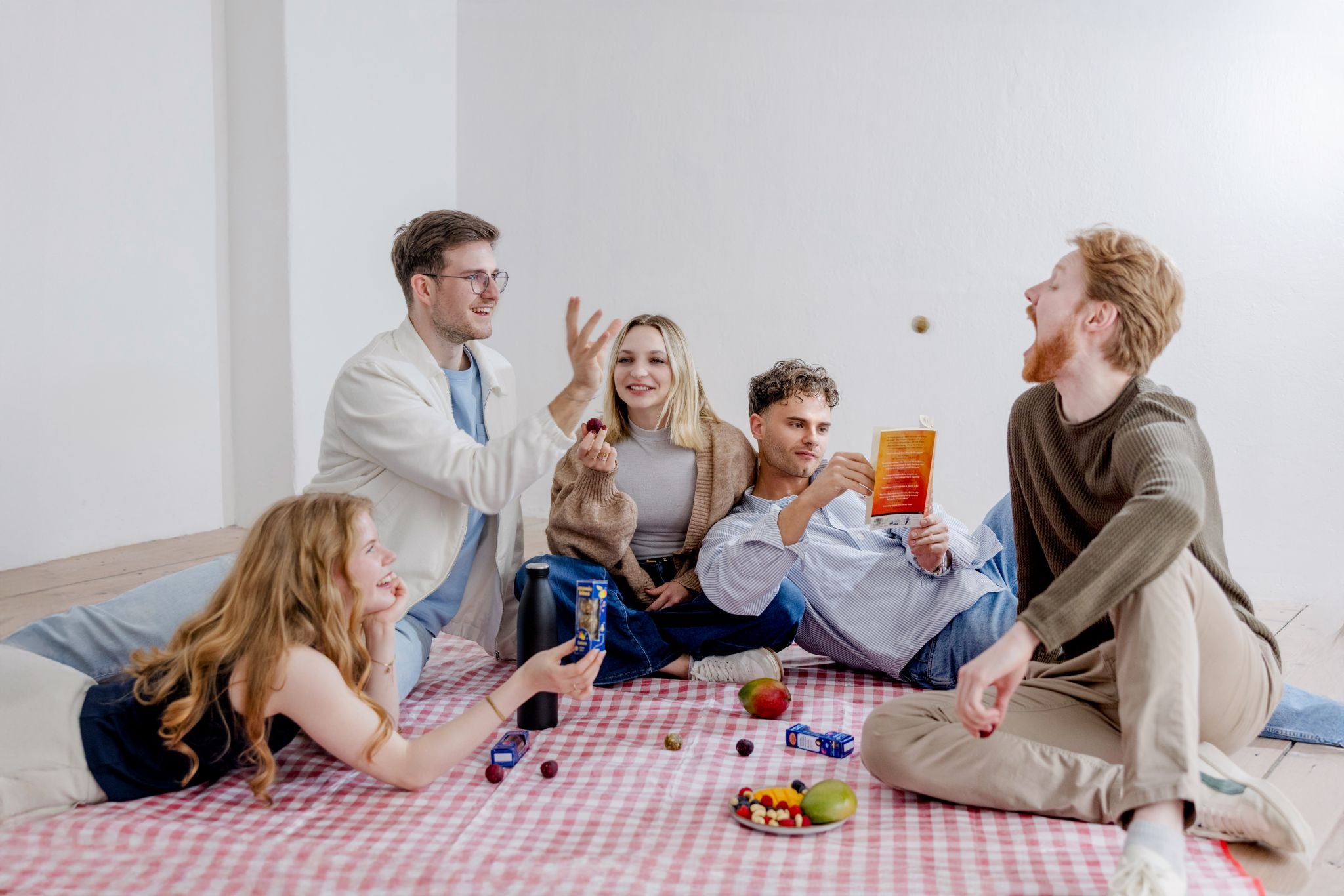 Eine junge Freundesgruppe genießt ein Picnic gemeinsam. Es wird gelesen, gelacht und Spaß gehabt mit Cosmic Bites Energy Balls, die hier nicht nur als gesunder Snack eingesetzt werden. Ein Freund wirft einen Cosmic Bites Energy Ball über die gesamte Picnic-Decke in den geöffneten Mund eines anderen Freundes. Verschiedene Packungen an Cosmic Bites Energy Balls liegen verteilt auf der Decke.
