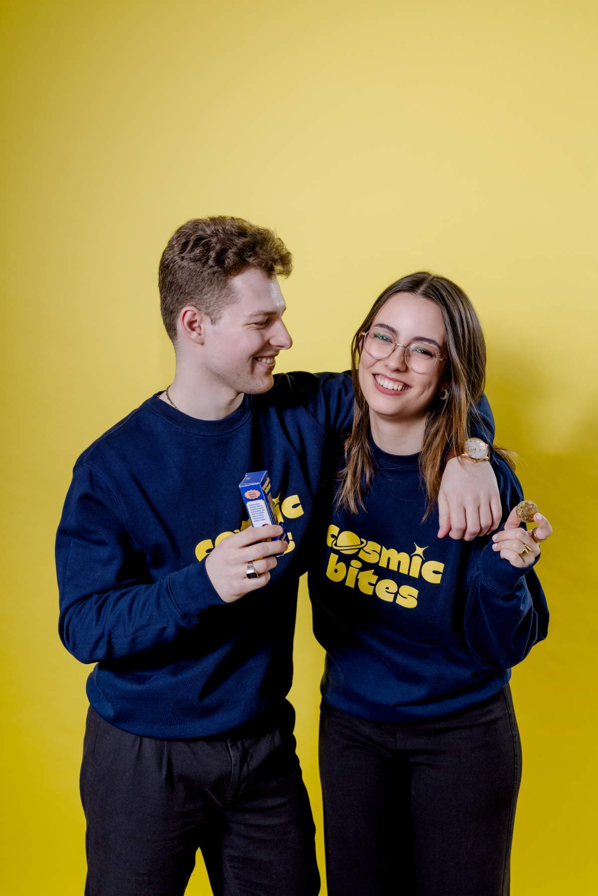 Die Gründer von Cosmic Bites Melanie Arndt und Laurin Mächtig liegen sich in den Armen. Sie tragen Pullis mit dem Logo ihrer Firma Cosmic Bites. Laurin hält eine Cosmic Bites Energy Ball Packung in der Hand, während Melanie einen einzelnen Energy Ball breit hält.
