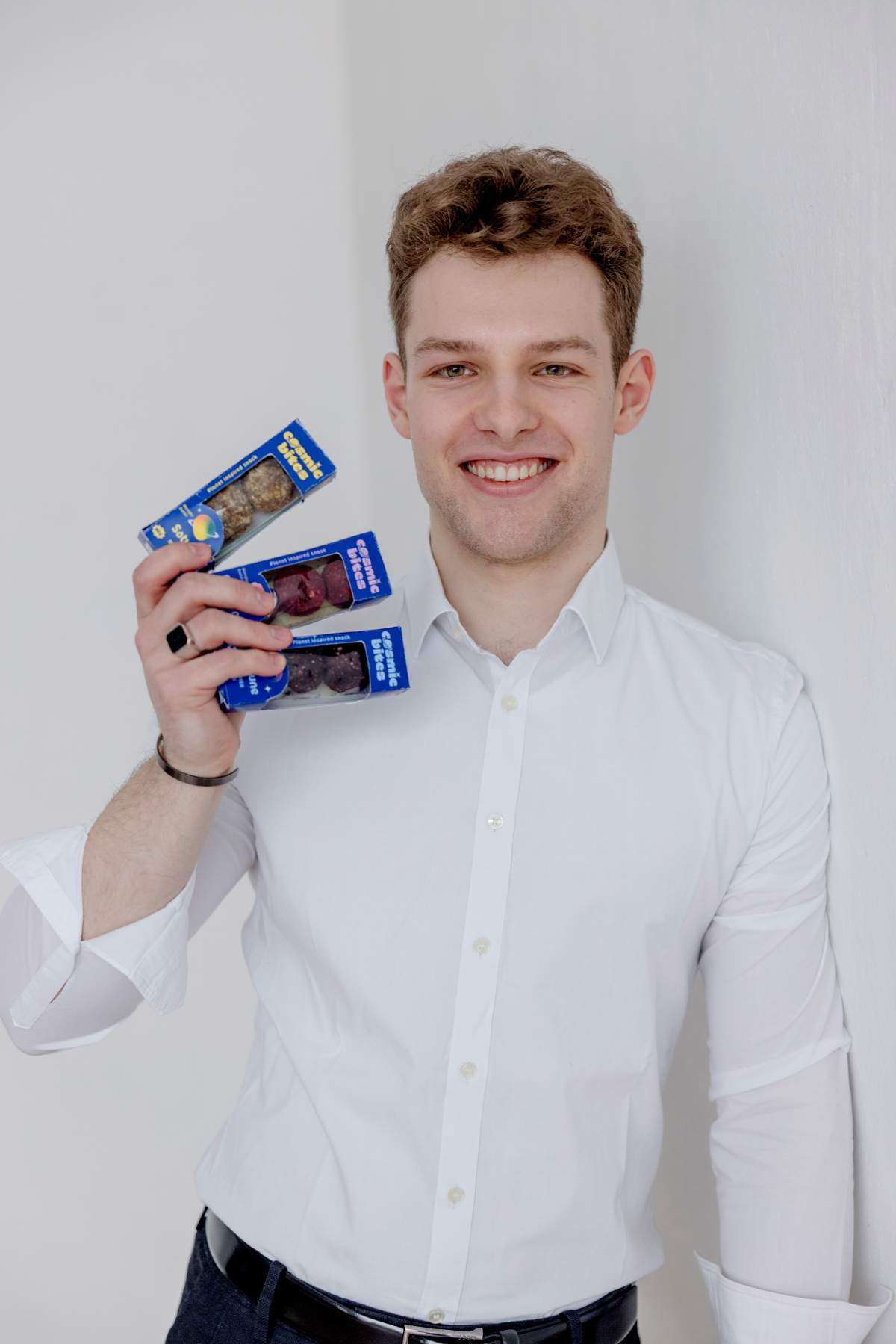 Der Gründer von Cosmic Bites Laurin Mächtig halt eine Packung von jeder der drei Sorten Cosmic Bites in die Kamera: Rote Energy Balls der Sorte Himbeere, die dem Planeten Mars nachempfunden sind, Gelbe Energy Balls der Sorte Mango, die dem Planeten Saturn nachempfunden sind, und tiefviolette Energy Balls der Sorte Heidelbeere, die dem Planeten Neptun nachempfunden sind.