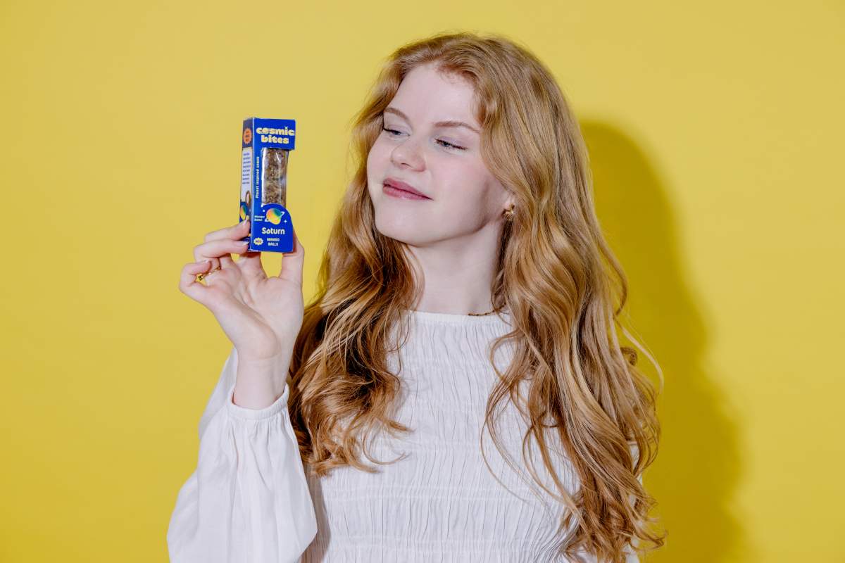 Eine junge Frau grinst und schaut auf eine Cosmic Bites Energy Ball Packung der Sorte Mango. Mit verträumten Blick schaut sie auf die gelben Cosmic Bites Energy Balls, die dem Planeten Saturn ähneln.