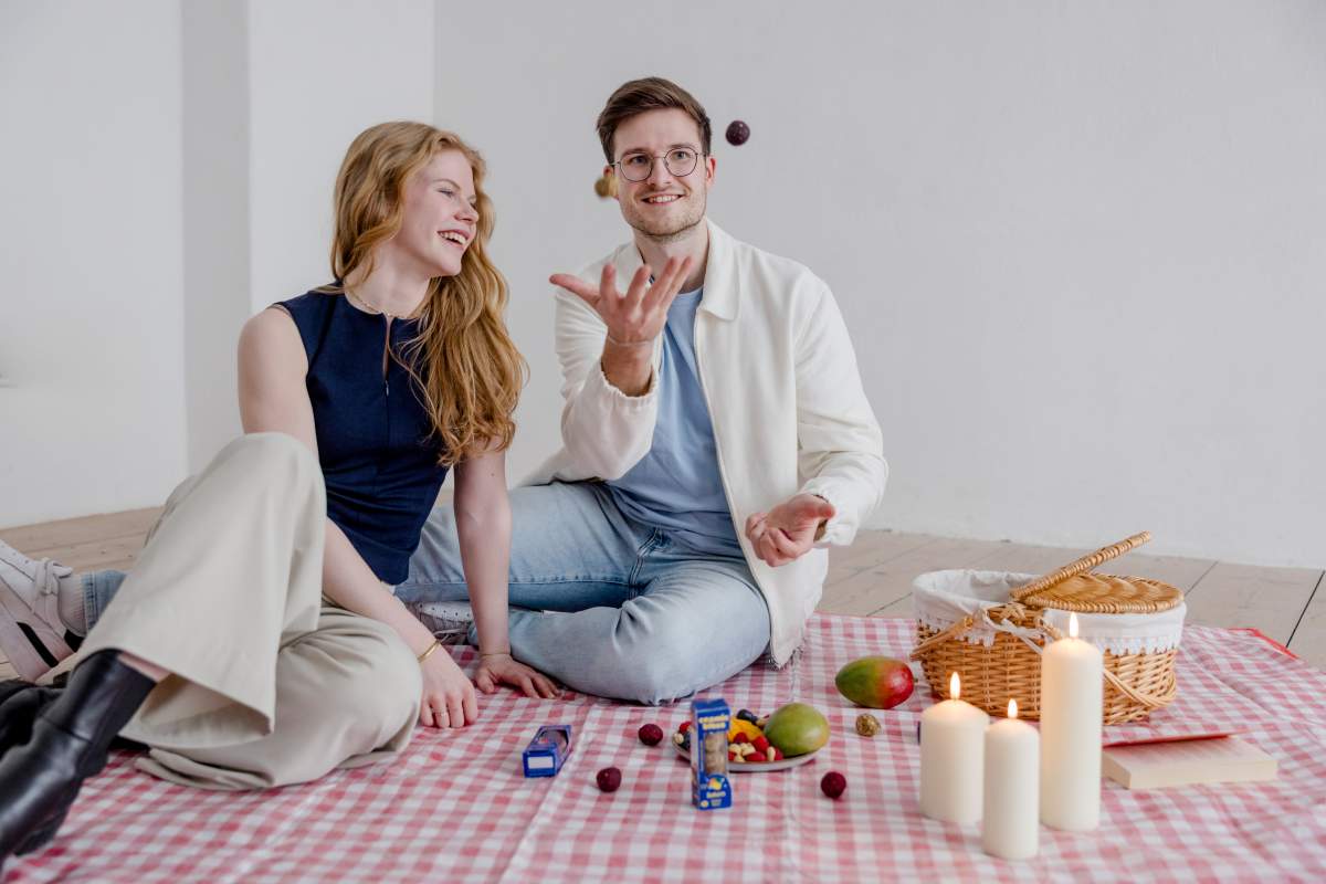Eine junge Frau und ein Mann sitzen auf einer Picnic-Decke im romantischen Kerzenschein. Der Mann jongliert ein paar Cosmic Bites Energy Balls, während sie lacht und den schwebenden gesunden Snacks zuschaut. Auf der Picnic-Decke liegen weitere Cosmic Bites Energy Ball Packungen, sowie einige Früchte und ein Picnic-Korb.