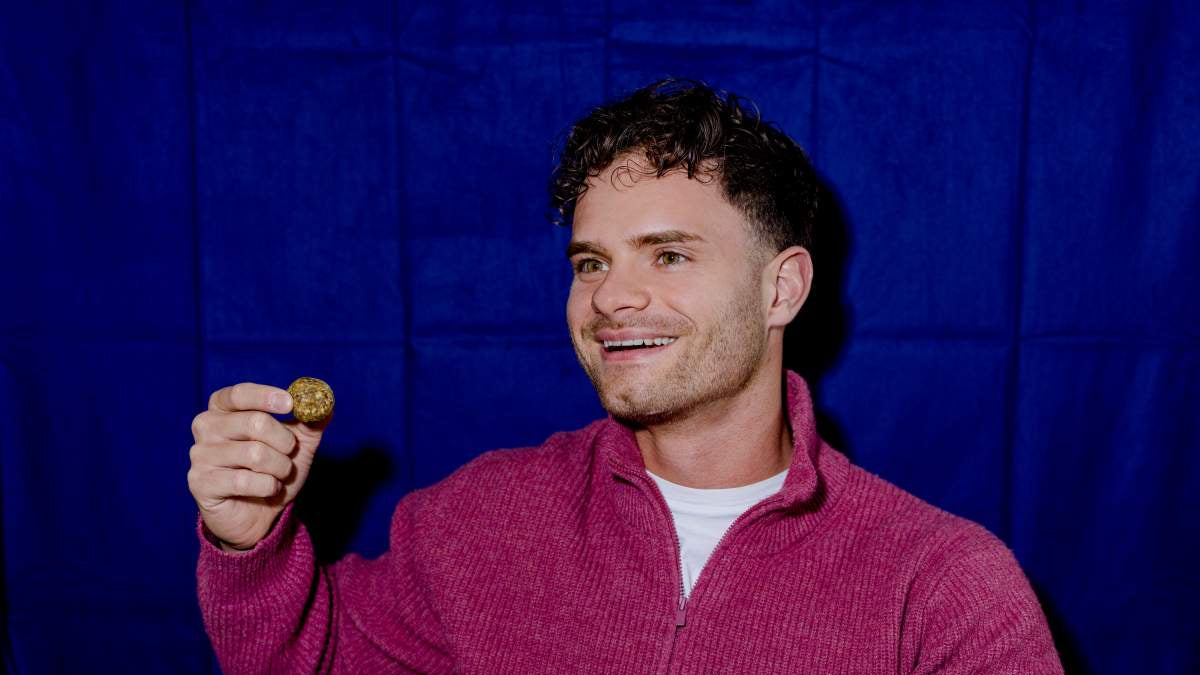 Ein junger lachender Mann hält einen Cosmic Bites Energy Ball der Geschmacksrichtung Mango in einer Hand. Vor dem dunkelblauen Hintergrund sieht es fast so aus, als würde der gesunde Snack schweben. Genauso, wie der Planet Saturn, dem dieser gelbe Energy Ball nachempfunden ist.