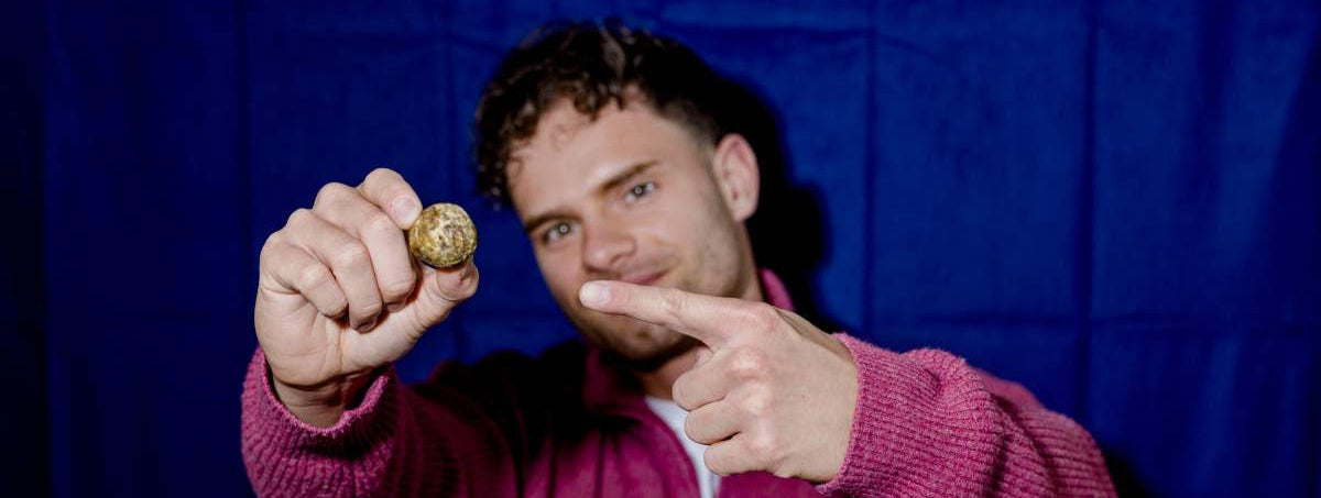 Ein junger Mann zeigt cool auf einen Cosmic Bites Energy Ball der Sorte Mango, welcher im Vordergrund geradezu heraussticht. Der gelbe Energy Ball ist nicht nur der gesunde Snack seiner Wahl, sondern sieht außerdem aus, wie der Planet Saturn.