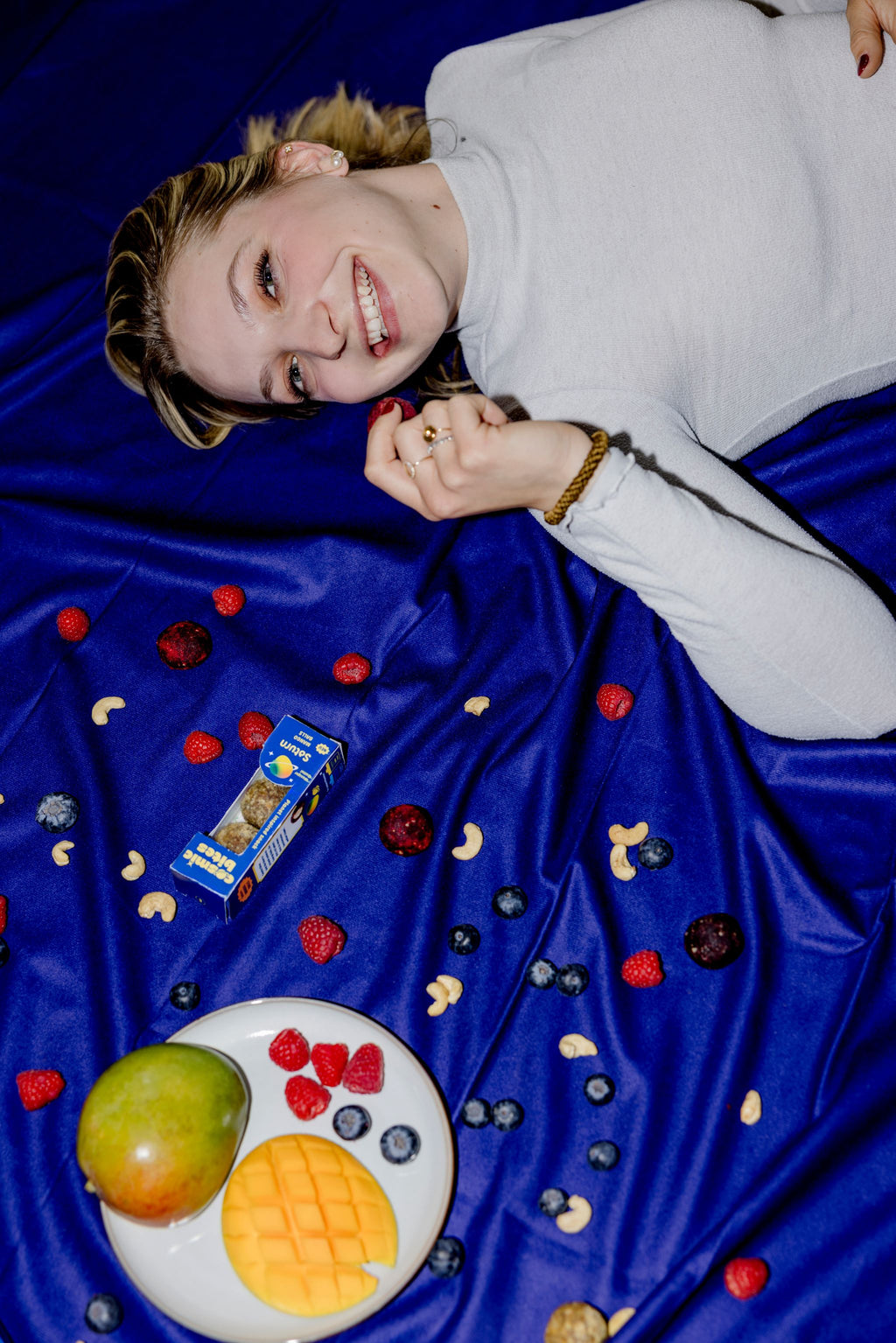 Eine junge Frau liegt auf dem Rücken auf einem tiefblauen Hintergrund. Es wirkt so, als würde sie entspannt durch die Tiefen des Alls schweben. Sie grinst und isst einen Cosmic Bites Energy Ball der Sorte Himbeere. Neben ihr schweben weitere Cosmic Bites Energy Balls durch das Weltall, sowie gesunde Zutaten wie Früchte und Cashew-Kerne, die auch in den gesunden Snacks enthalten sind.