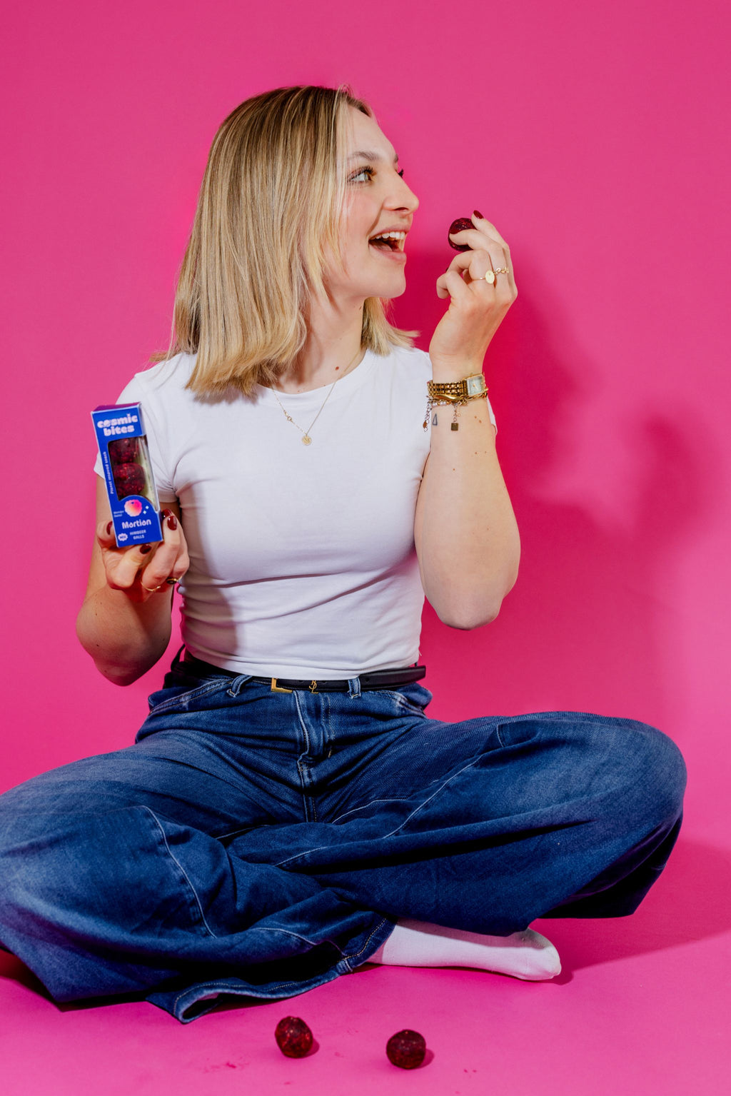 Eine junge Frau sitzt im T-Shirt im Schneidersitz auf dem Boden und genießt einen Cosmic Bites Energy Ball der Sorte Himbeere. In ihrer anderen Hand hält sie schon die nächste Packung parat. Die strahlend roten Energy Balls stehen im Fokus. Ein paar weitere Bälle liegen vor ihr und sehen aus, wie kleine Versionen des Planeten Mars.