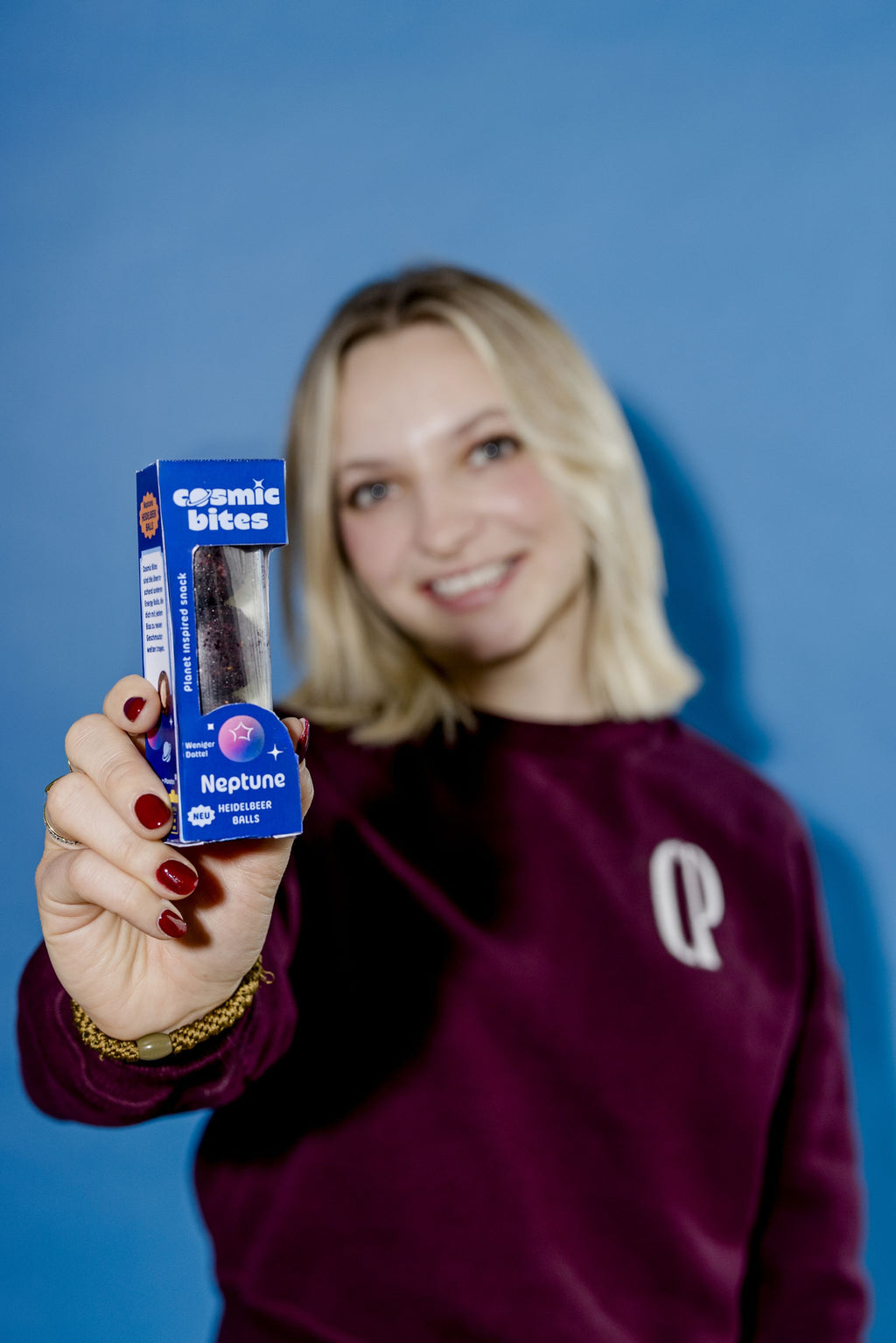Eine junge Frau hält eine Cosmic Bites Energy Ball Packung der Sorte Heidelbeere in den Vordergrund, während sie leicht im Hintergrund verschwindet und lächelt. Die tiefvioletten Bälle in der Packung stechen hervor und sehen aus, wie kleine Abbilder des Planeten Neptun.