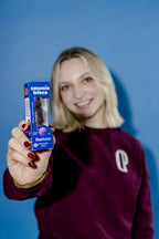 Eine junge Frau hält eine Cosmic Bites Energy Ball Packung der Sorte Heidelbeere in den Vordergrund, während sie leicht im Hintergrund verschwindet und lächelt. Die tiefvioletten Bälle in der Packung stechen hervor und sehen aus, wie kleine Abbilder des Planeten Neptun.