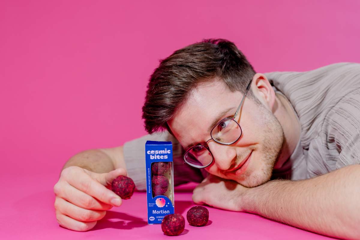 Ein junger Mann liegt auf dem Bauch und schaut zufrieden und glücklich auf eine Packung Cosmic Bites Energy Balls der Sorte Himbeere. Daneben liegen zwei weitere Himbeer Energy Balls und er hat einen Himbeer Energy Ball in der Hand. Die Cosmic Bites Energy Balls sind strahlend rot und sehen aus wie kleine Versionen des Planeten Mars.