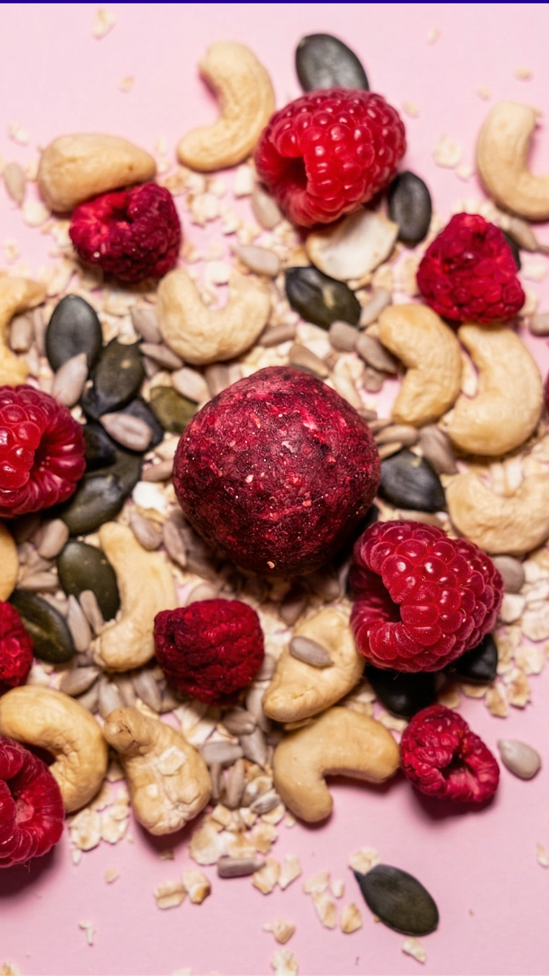Knallig roter Cosmic Bites Energy Ball inspiriert vom Planeten Mars, umgeben von frischen Himbeeren, Cashews und Kernen – leckerer gesunder Snack mit wenig Datteln.